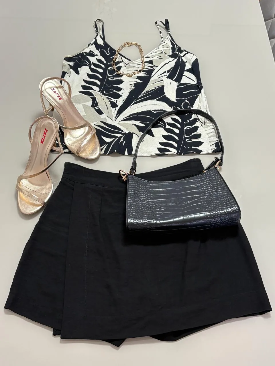 Look com regata estampada de folhagens, saia preta, bolsa croco e sandália dourada montado sobre uma mesa